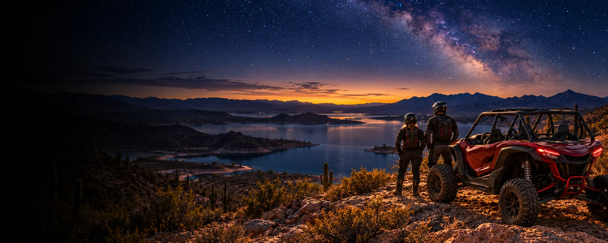 Two riders in helmets beside a red side-by-side on a rocky ridge at dusk, with the Milky Way rising over a desert lake and saguaros in the foreground